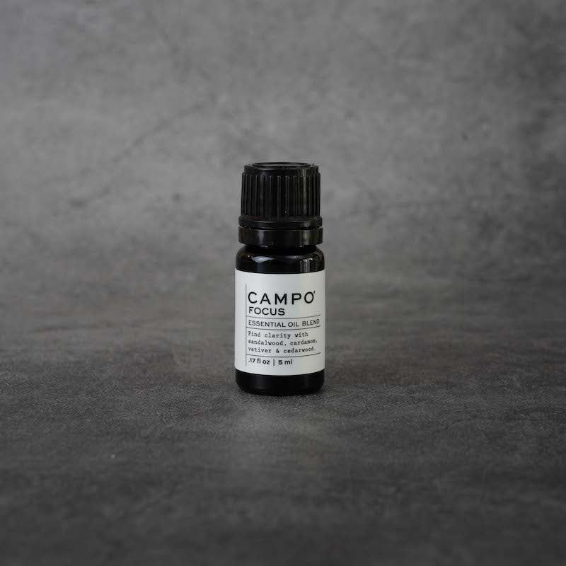 A small black bottle with a white label. The label reads "CAMPO Focus Essential oil blend, Find clarity with sandalwood, cardamom, vetiver & cedarwood , .17 fl oz, 5 ml" in black text. The bottle has a black twist-off lid.