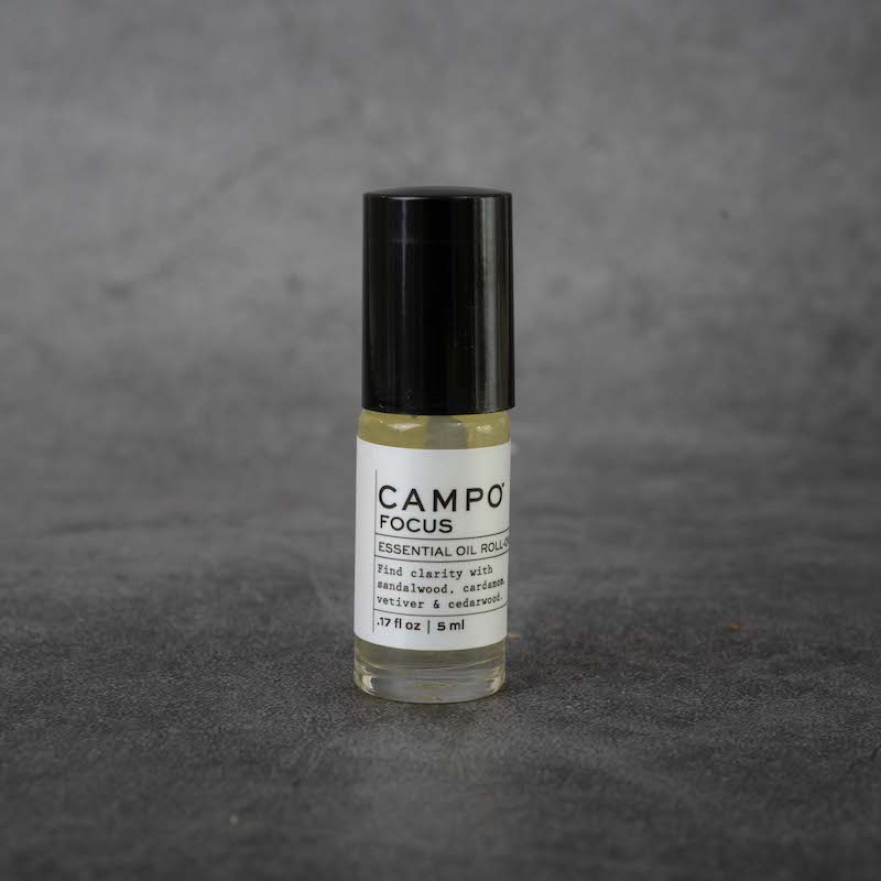 A small clear glass bottle with a white label. The label reads "CAMPO Focus Essential Oil Roll-on, Find clarity with sandalwood, cardamom, vetiver & cedarwood, .17 fl oz, 5 ml". The bottle has a black twist-off lid.