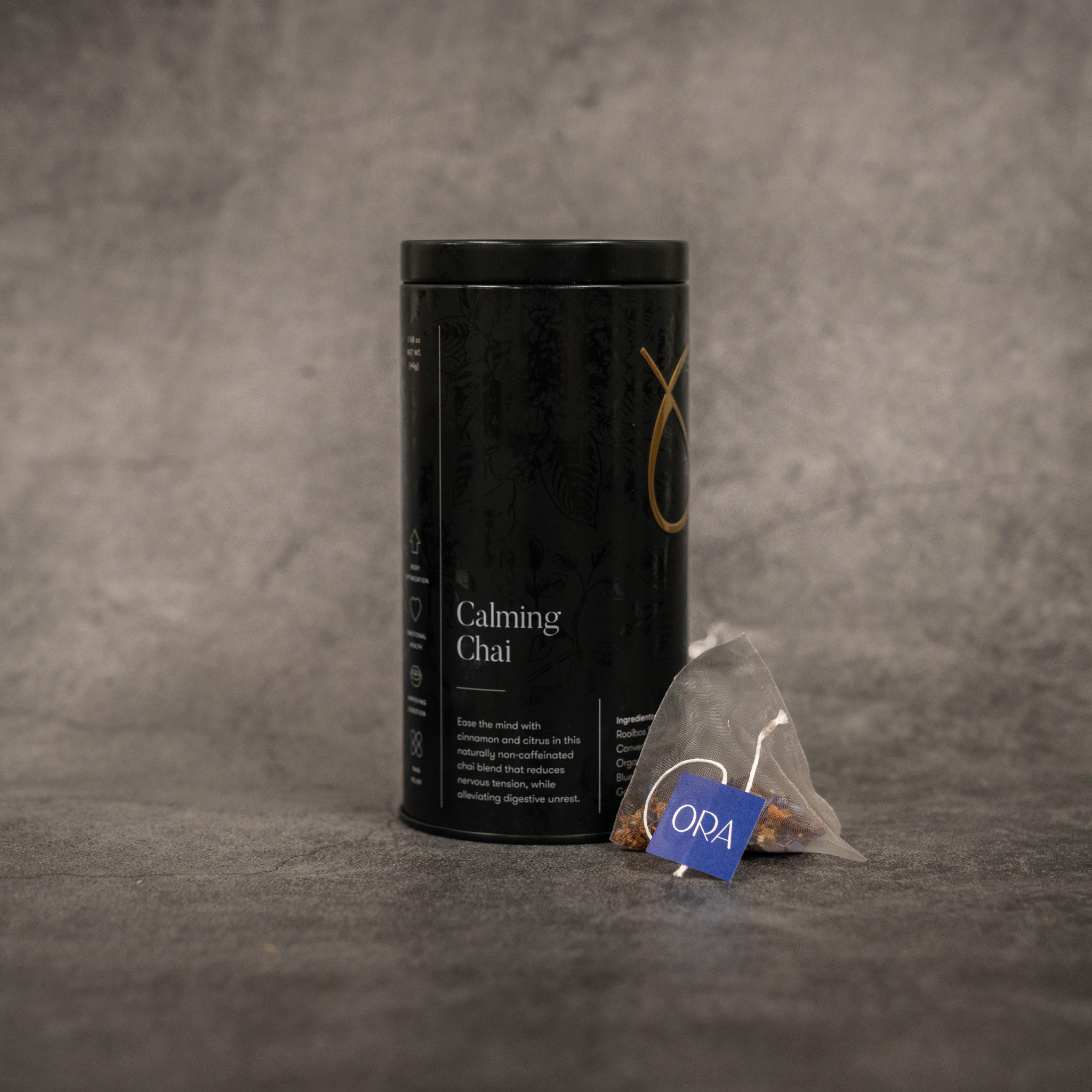 On the left, a black metal cylinder tin of tea reading "Calming Chai". On the right, a translucent tea bag with a string printed with the ORA logo. 