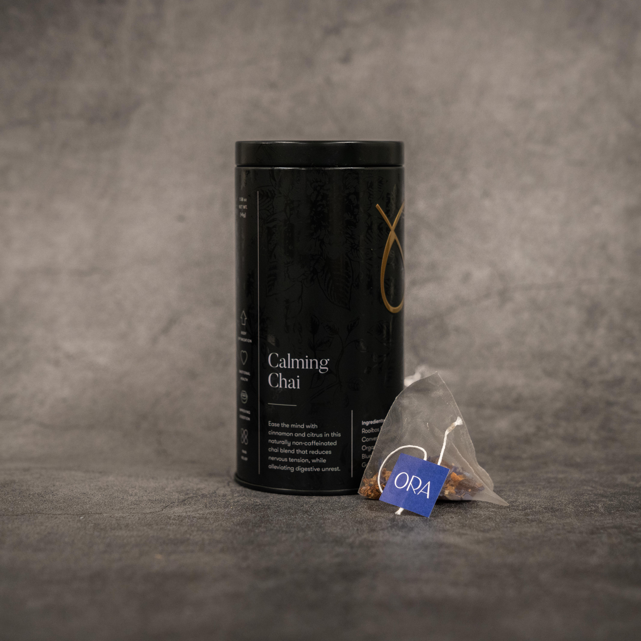 On the left, a black metal cylinder tin of tea reading "Calming Chai". On the right, a translucent tea bag with a string printed with the ORA logo.