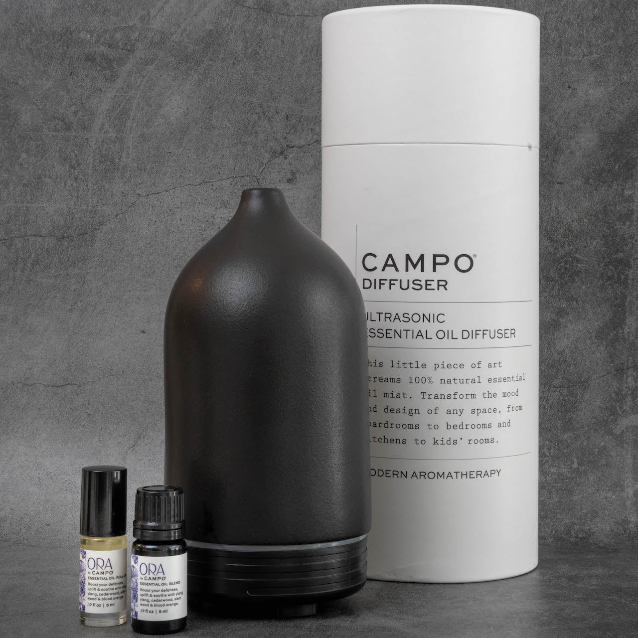 The same black diffuser as in the previous picture, with a set of ORA by Campo essential oils next to it. Behind the diffuser is the packaging for the diffuser, a tall white cylinder reading "Campo Diffuser, Ultrasonic Essential Oil Diffuser".