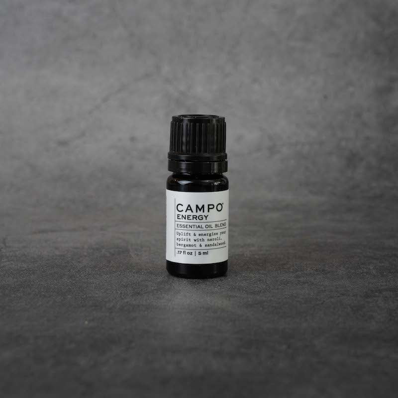 A small black bottle with a white label. The label reads "CAMPO Energy Essential oil blend, Uplift & energize your spirit with neroli, bergamot & sandalwood, .17 fl oz, 5 ml" in black text. The bottle has a black twist-off lid.