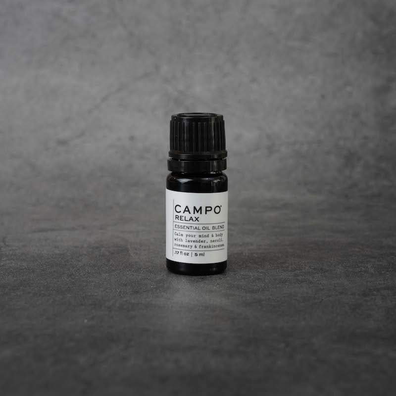 A small black bottle with a white label. The label reads "CAMPO Relax Essential oil blend, Calm your mind & body with lavender, neroli, rosemary & frankincense, .17 fl oz, 5 ml" in black text. The bottle has a black twist-off lid.