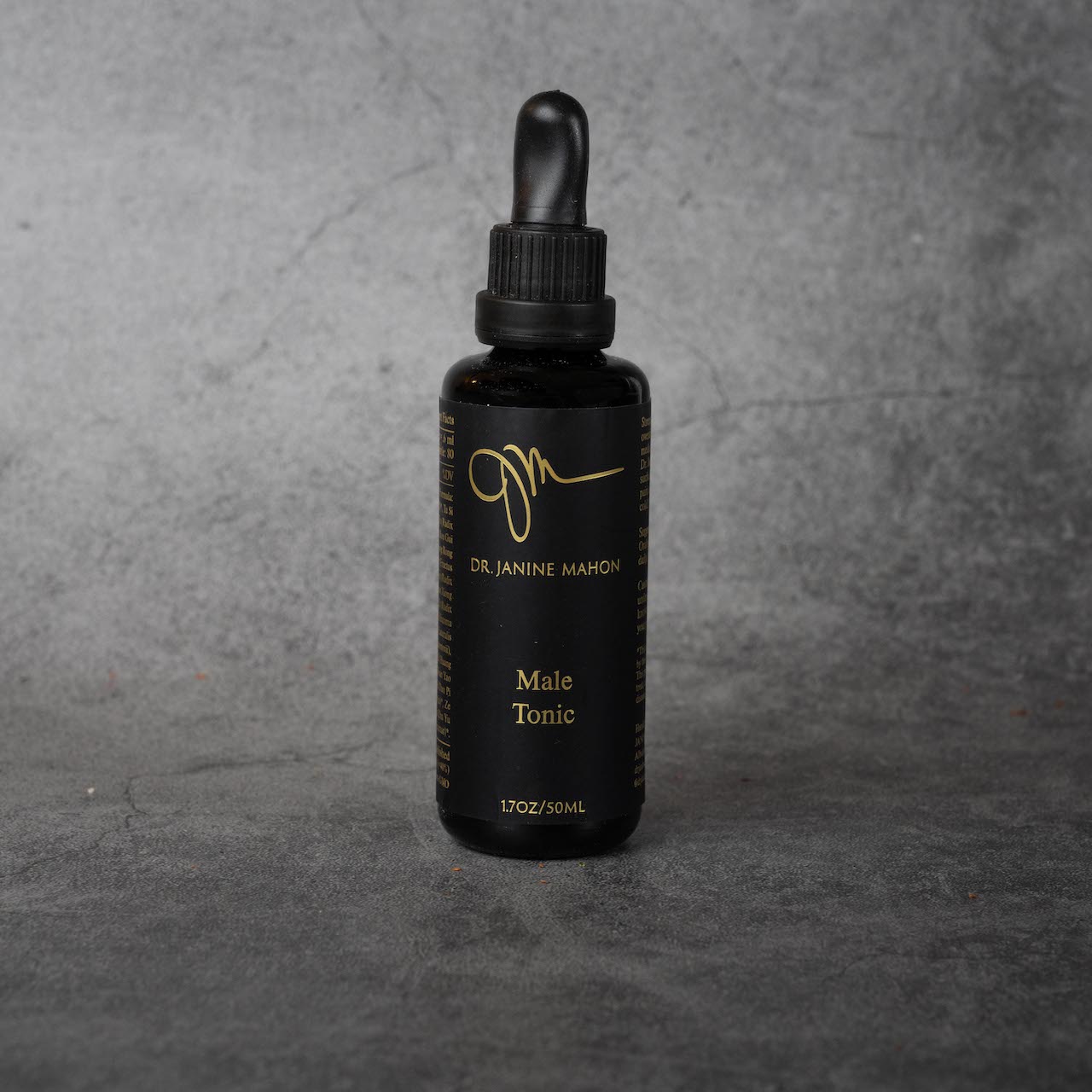 A small black bottle with gold lettering reading "Dr. Janine Mahon" in small print. Below, in slightly larger print, "Male Tonic, 1.7oz/50ML". The bottle has a twist-off lid and a silicone dropper.