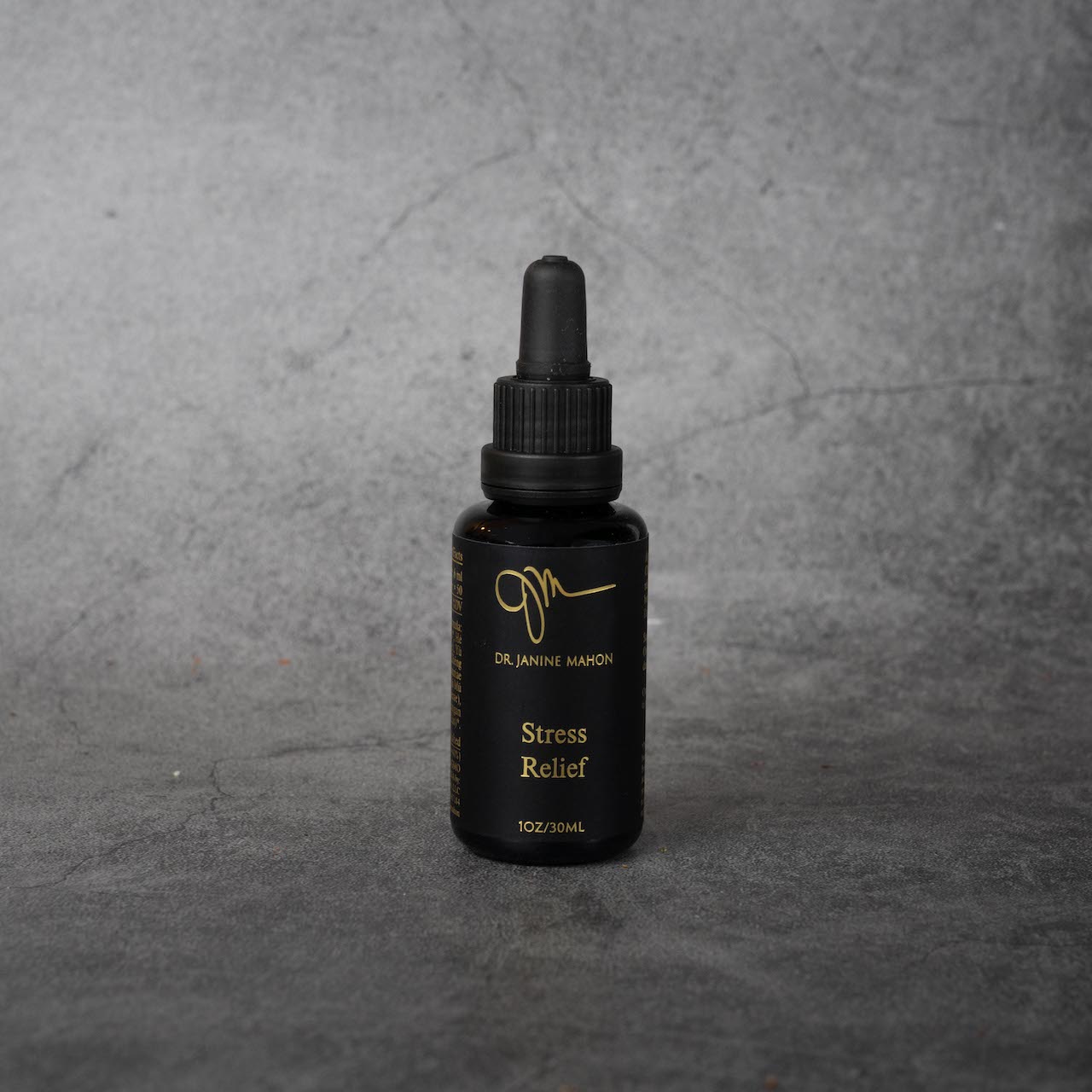 A small black bottle with gold lettering reading "Dr. Janine Mahon" in small print, and "Stress Relief, 1oz/30ml" in slightly larger print. The bottle has a twist-off silicone dropper top.