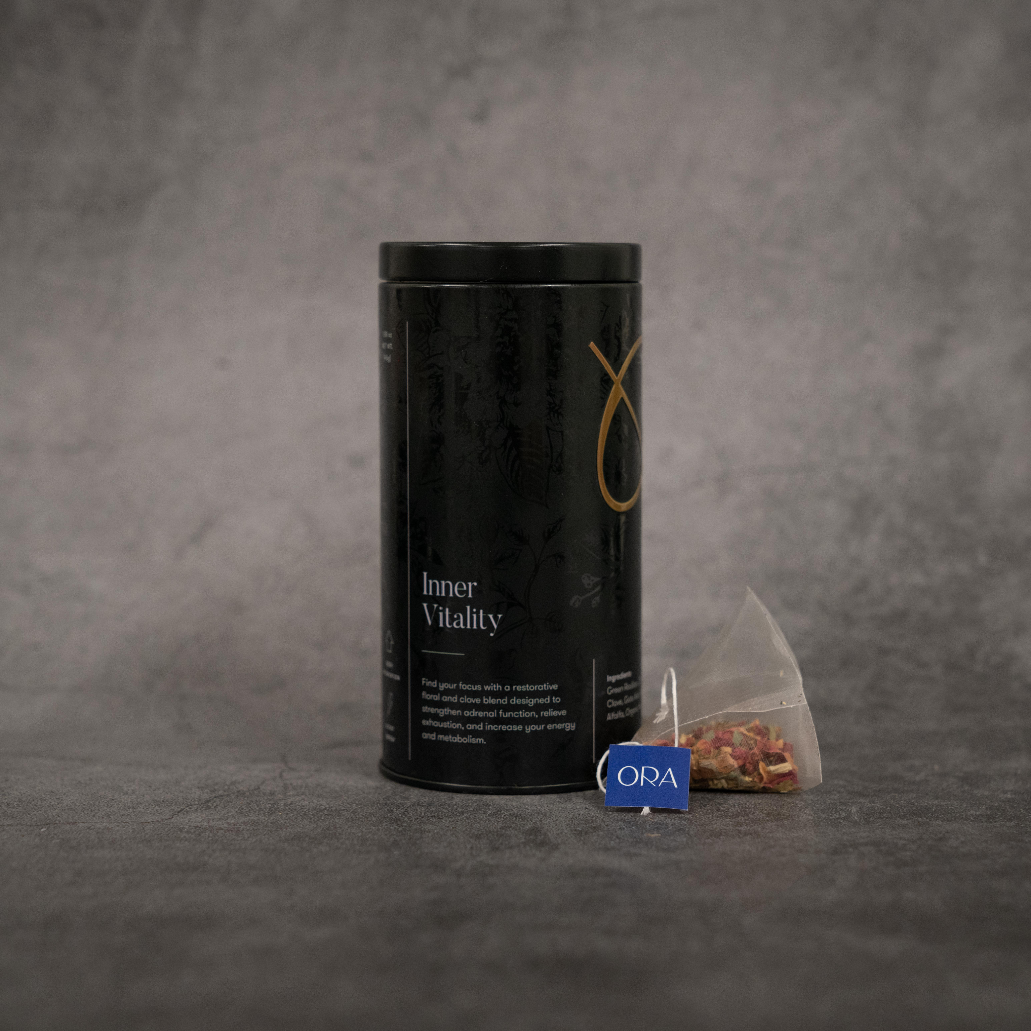 On the left, a black metal cylinder tin of tea reading "Inner Vitality". On the right, a translucent tea bag with a string printed with the ORA logo.