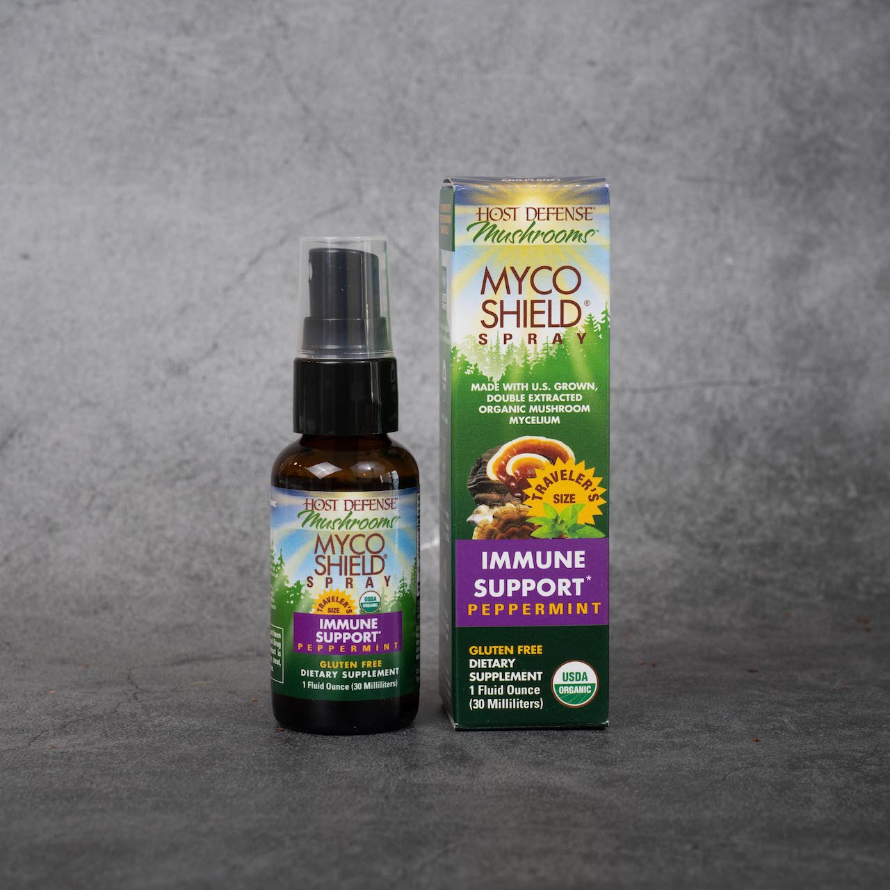 On the left, a small spray bottle. The label on the bottle has a drawing of trees and a sunny sky, and reads "Host Defense Mushrooms, Myco Shield Spray, Traveler's Size, Immune Support Peppermint, Gluten Free Dietary Supplement, 1fl oz, 30ml". It has a USDA organic certification label. On the right, the box for the product. It has the same background drawing, plus illustrations of mushrooms. It has the same text as the bottle, plus white text reading "Made with U.S. grown, double extracted organic mushroom 