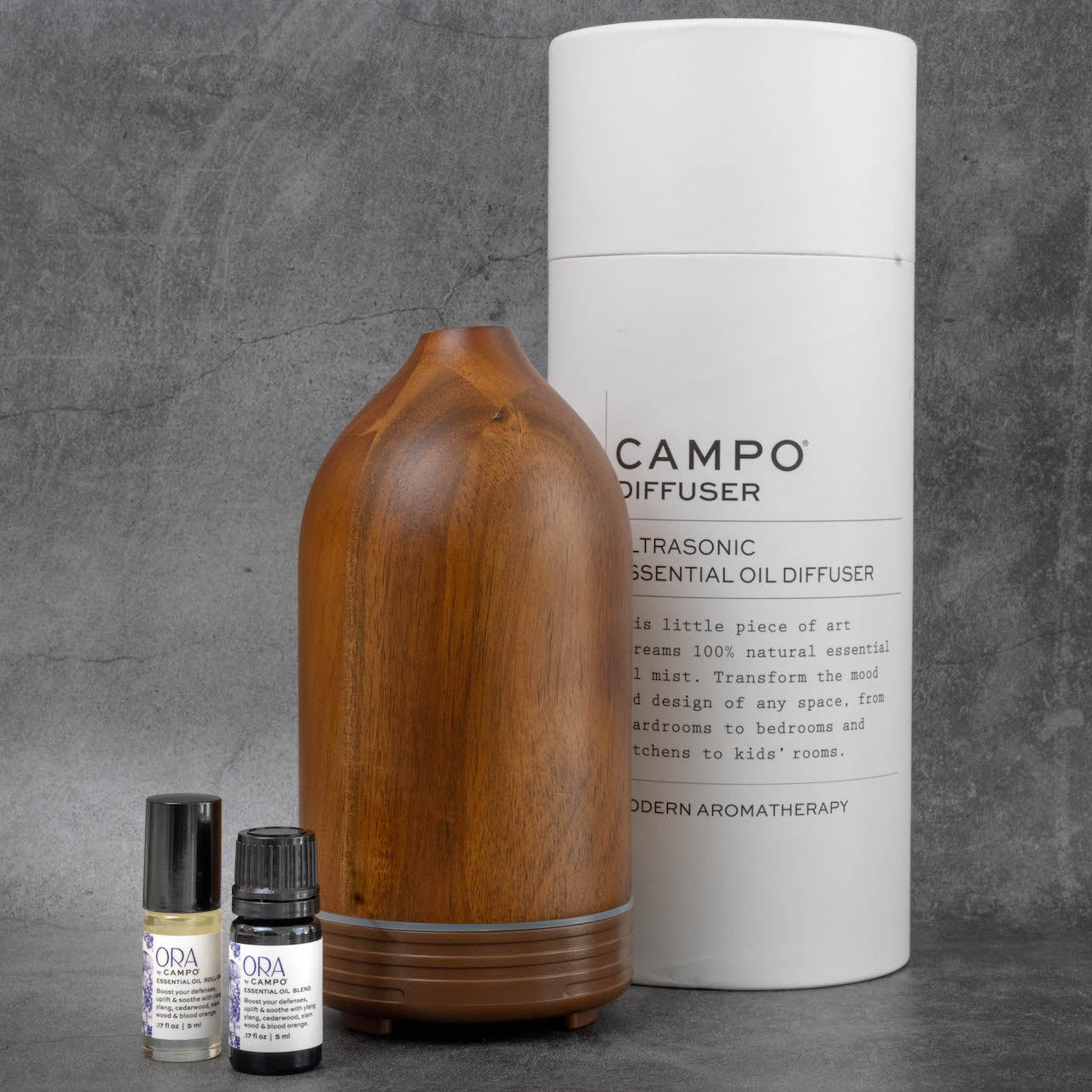 The same wooden diffuser as in the previous picture, with a set of ORA by Campo essential oils next to it. Behind the diffuser is the packaging for the diffuser, a tall white cylinder reading "Campo Diffuser, Ultrasonic Essential Oil Diffuser".
