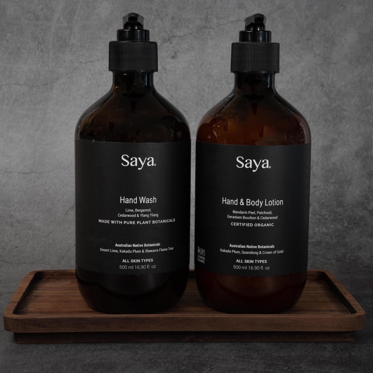 Two large, dark colored glass bottles sitting next to each other, each with a black pump-top and a black label. The white lettering on the bottle on the left reads " Saya Hand Wash, Lime, Bergamot, Cedarwood, & Ylang Ylang, Made with pure plant botanicals, Australian Native Botanicals, Desert Lime, Kakadu Plum & Illawarra Flame Tree, All skin types, 500ml/16.9 fl oz". The white lettering on the bottle on the right reads, "Saya Hand & Body Lotion, Mandarin Peel, Patchouli, Geranium Bourbon, & Cedarwood, Cert