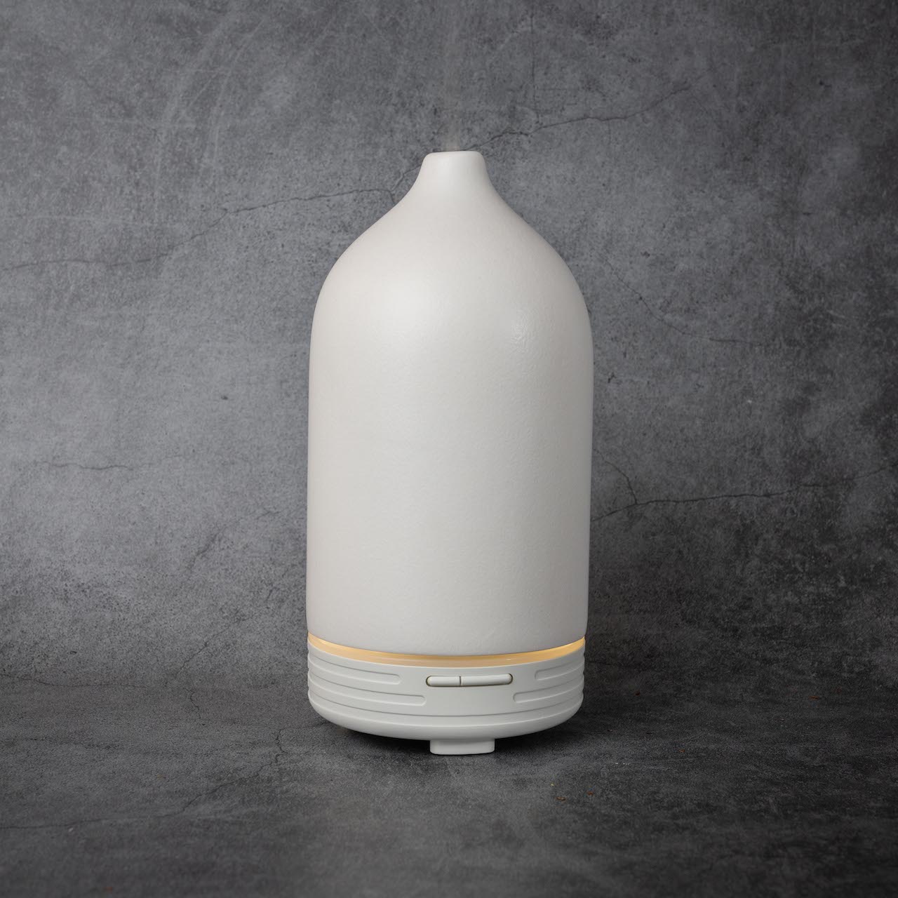 The same white, cone shaped diffuser as in the previous photo, now with power on. There are two small oval shaped buttons near the base of the diffuser, with a thin band above them that is lit up.